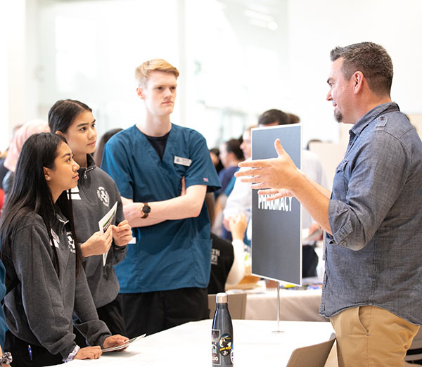 Three young people speak to man at a tabling event. Man holds up his hands to gesture.