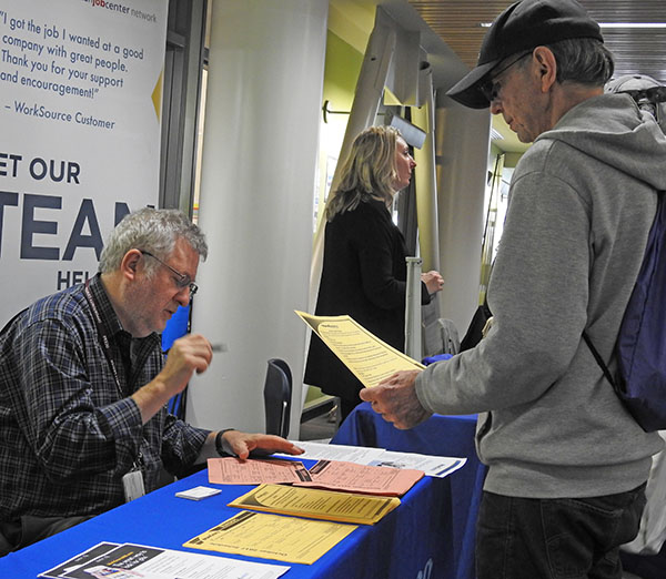 Man in baseball hat holds up paper while listening to a man behind a table with stacks of papers.